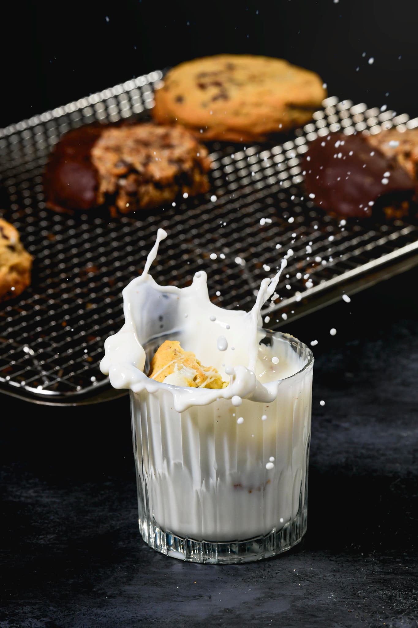 A Claudette's cookie dropping into a glass of milk, fresh batch cooling behind